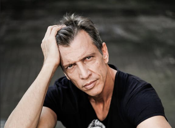 Portrait de Rémi Desrochers, vêtu d’un t-shirt noir, la main dans les cheveux et le regard sérieux.